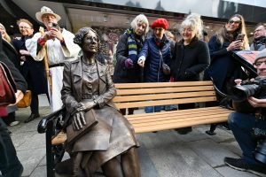 Bronze statue of Townsend Warner sitting at the end of a wooden bench, surrounded by people pointing and taking photos